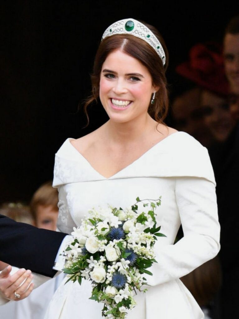 Bride with brown hair holds bouquet of white flowers, wearing a white gown and a silver tiara set with a large emerald