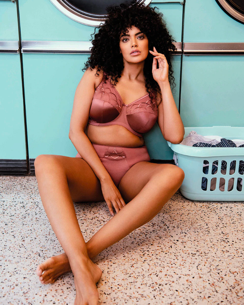 A woman sits on the ground against teal washing machines resting an elbow on a washing basket, wearing rose gold matching underwear and bra