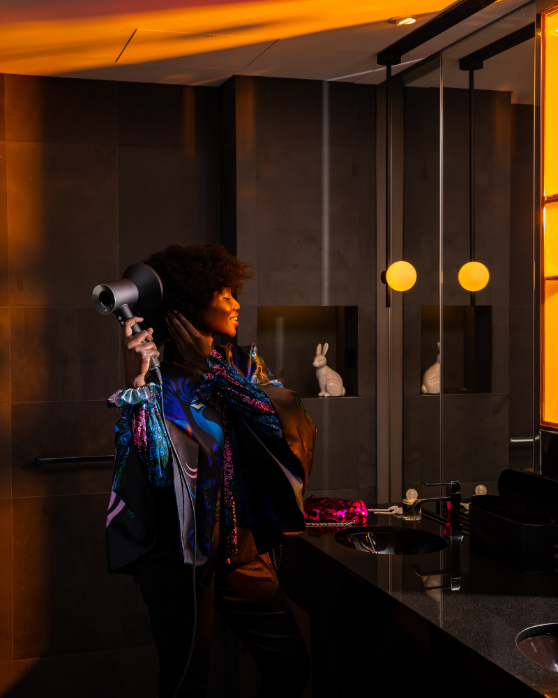 A woman in a hotel bathroom in dim lighting, blow drying her curly hair
